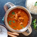 An overhead shot of tikka chicken masala soup in a pot with empty bowls beside it with another bowl of rice.