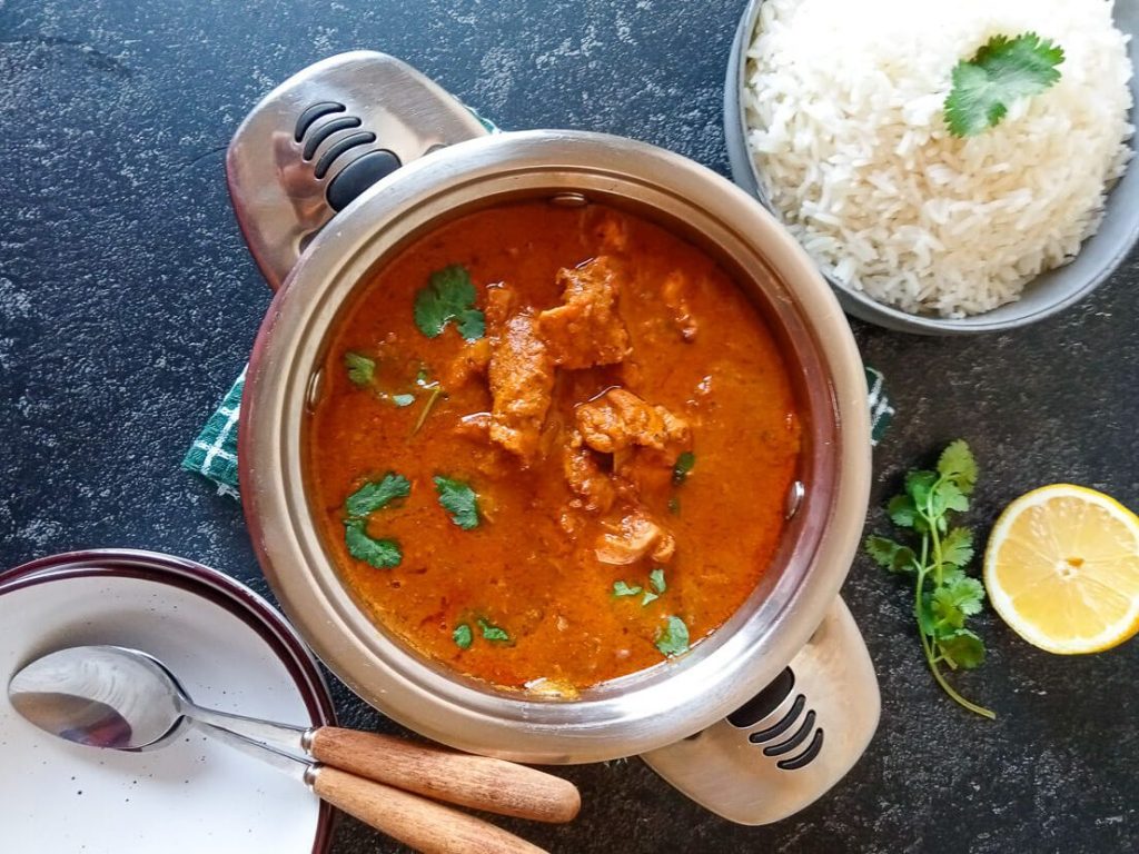 An overhead shot of tikka chicken masala soup in a pot with empty bowls beside it with another bowl of rice.