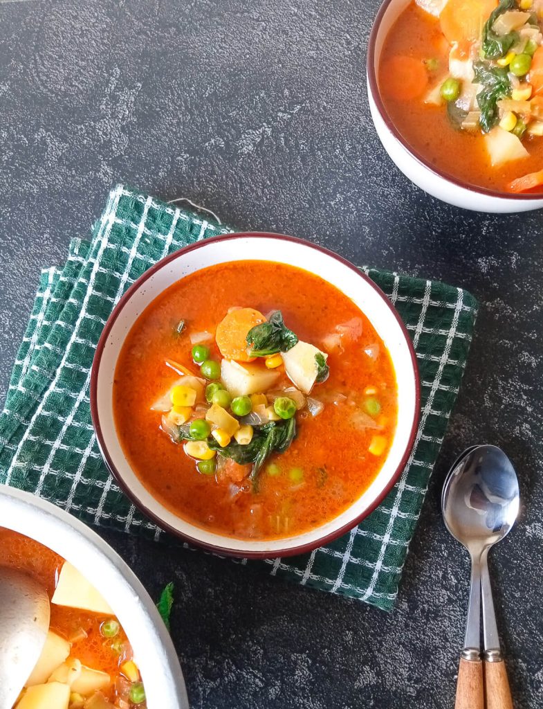 An image of bone broth vegetable soup in bowls with one on top of a green tablecloth with two spoons beside it