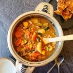 A pot of barley minestrone soup with empty serving bowls next to it. There is also a black plate with toasted bread in it all this is on a black background.