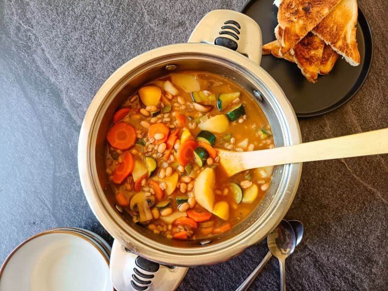 A pot of barley minestrone soup with empty serving bowls next to it. There is also a black plate with toasted bread in it all this is on a black background.