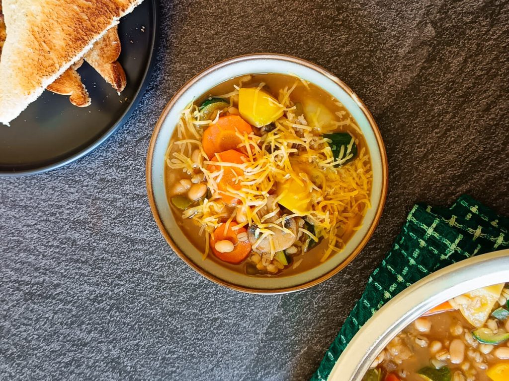 A cheesy bowl of barley minestrone soup with a pot and a black plate with toasted bread in it next to it. All this is on a black background.