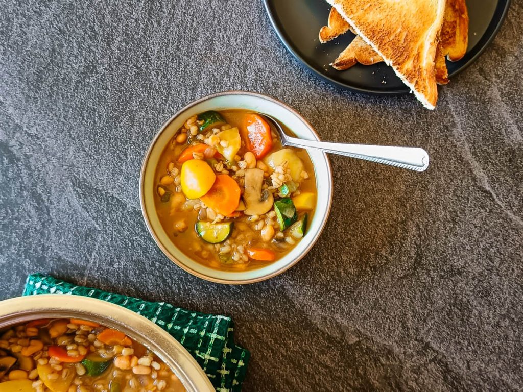 A bowl of barley minestrone soup with a pot and a black plate with toasted bread in it next to it. All this is on a black background.