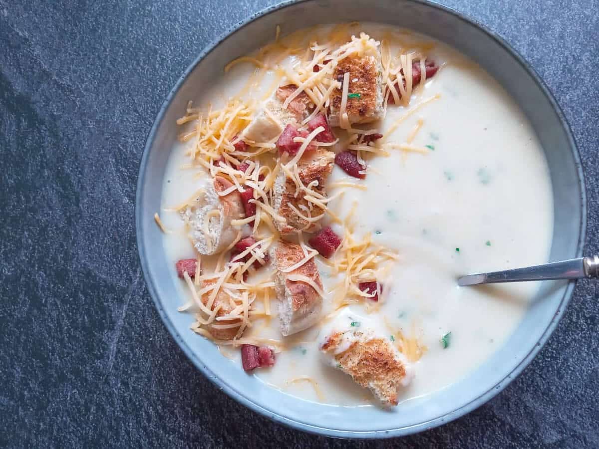 Garlic soup topped with croutons, fried spam, and shredded cheese in bowl with black background.