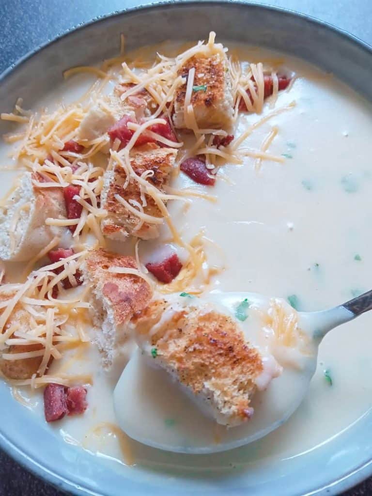 Garlic soup topped with croutons, fried spam, and shredded cheese in bowl with black background.