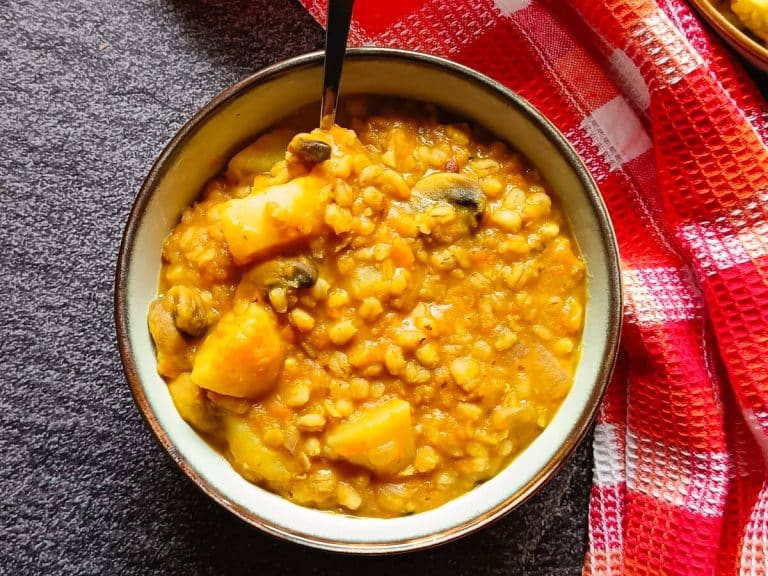 A bowl of Barley and Lentil soup with a silver and wooden spoon with a tablecloth near it on a black background.