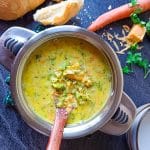 A pot of cheddar broccoli soup with a wooden spoon in it and two empty bowls near it with bread and other leftover ingredients
