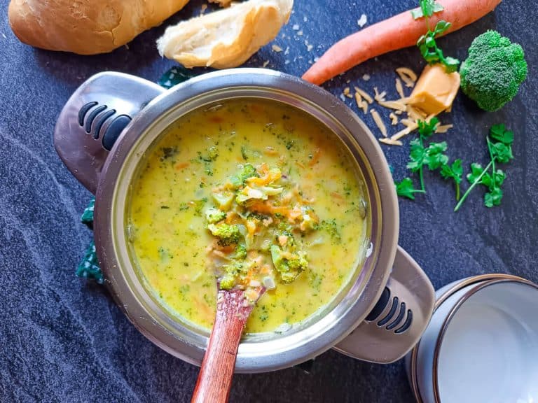 A pot of cheddar broccoli soup with a wooden spoon in it and two empty bowls near it with bread and other leftover ingredients