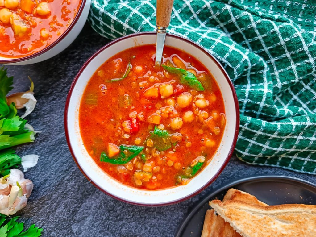 An overhead image of lentil chickpea soup bowl with a snap of a plate with toasted bread nearby and some leftover ingredients on a black background