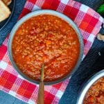 A bowl of red lentil tomato soup with a spoon in it on a tablecloth with a black background