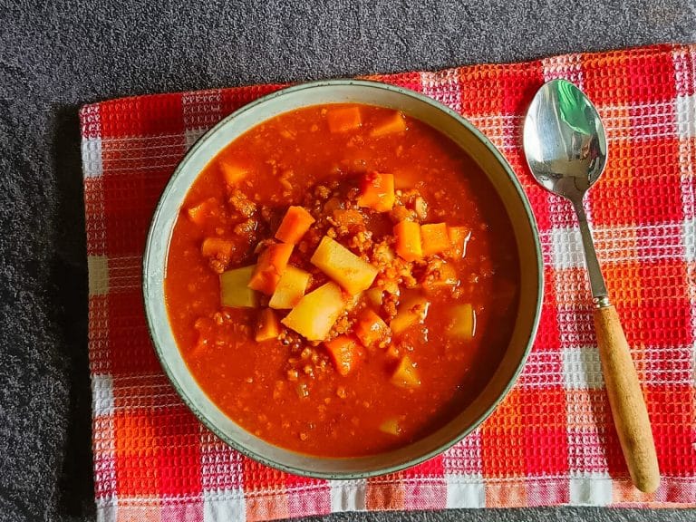 An overshot of a Mexican Picadillo Soup bowl with a spoon beside it on an orange and red tablecloth, all this on a black background.