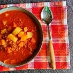 An overshot of a Mexican Picadillo Soup bowl with a spoon beside it on an orange and red tablecloth, all this on a black background.
