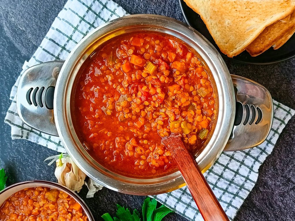 A pot of Moroccan lentil soup with a wooden spoon in it on a white and green tablecloth. There are some garlic skin and garlic cloves near the pot and another bowl of lentil soup with a plate of toasted bread beside it too, all this on a black background.