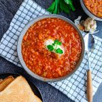A bowl of Moroccan lentil soup with a dollop of cream on a white and green tablecloth with a spoon beside it. There are some garlic skin and garlic cloves near the bowl and another bowl of lentil soup with a plate of toasted bread beside it too, all this on a black background.