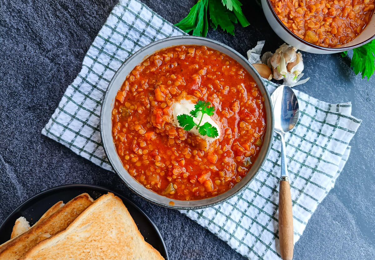 A bowl of Moroccan lentil soup with a dollop of cream on a white and green tablecloth with a spoon beside it. There are some garlic skin and garlic cloves near the bowl and another bowl of lentil soup with a plate of toasted bread beside it too, all this on a black background.