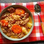A bowl of prime rib and barley soup on an orange tablecloth with a spoon beside it. All this is on a black background.