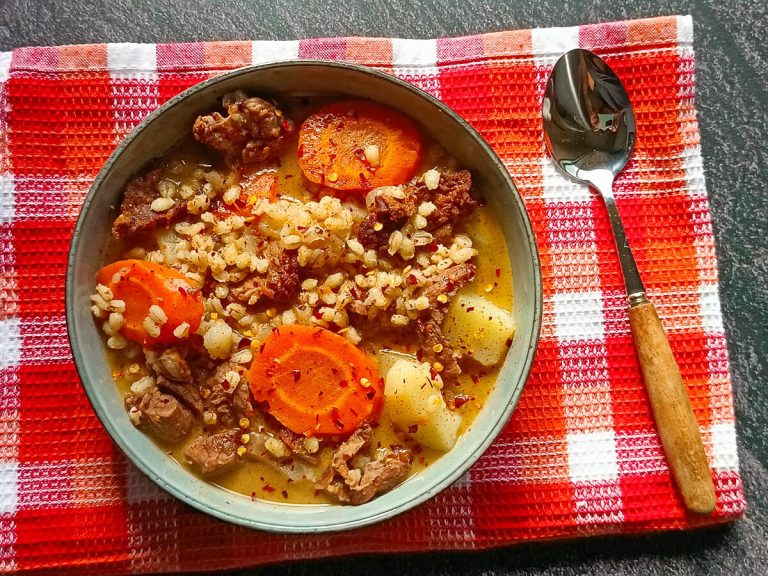 A bowl of prime rib and barley soup on an orange tablecloth with a spoon beside it. All this is on a black background.