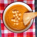 A bowl of tomato chickpea soup on a red and orange tablecloth on a black background.