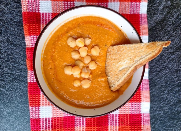 A bowl of tomato chickpea soup on a red and orange tablecloth on a black background.