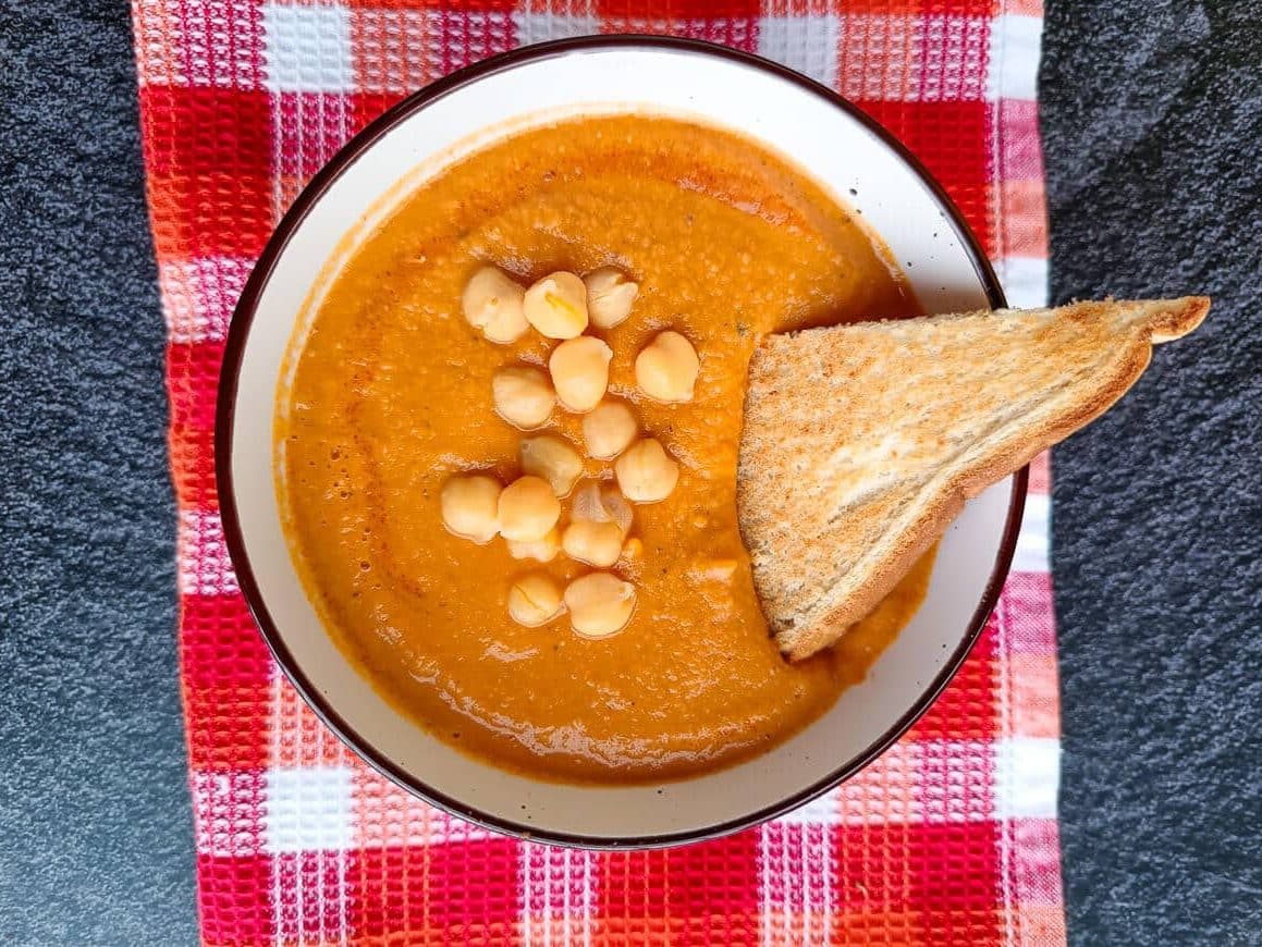 A bowl of tomato chickpea soup on a red and orange tablecloth on a black background.