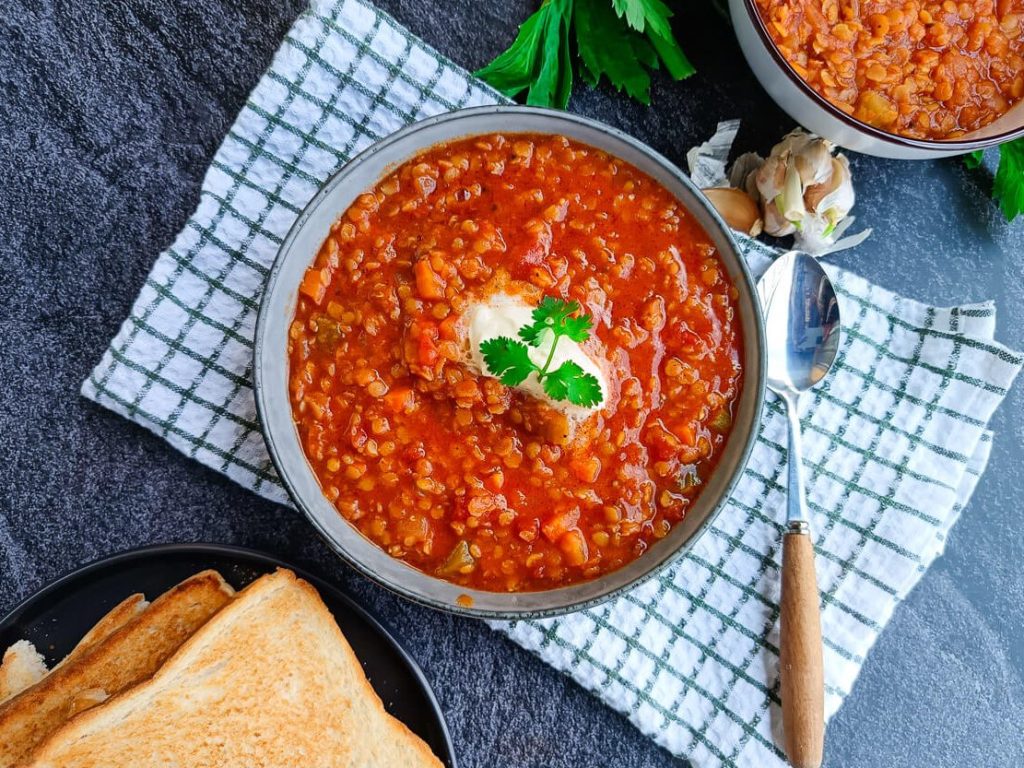 A bowl of Moroccan lentil soup with a dollop of cream on a white and green tablecloth with a spoon beside it. There are some garlic skin and garlic cloves near the bowl and another bowl of lentil soup with a plate of toasted bread beside it too, all this on a black background.