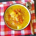 A bowl of potato and carrot soup sprinkled with chili flakes on a red and white tablecloth, with a silver spoon beside it on a black background.