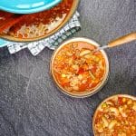 Two bowls and a blue pot of Busy Day Soup on a black background with a tablecloth. The hearty soup is made with elbow macaroni, ground beef, canned tomatoes, onions, celery, mixed vegetables, and beef stock. The hero bowl in the center has a spoon, showcasing the comforting, easy Busy Day Soup recipe.