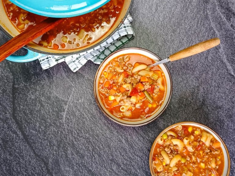 Two bowls and a blue pot of Busy Day Soup on a black background with a tablecloth. The hearty soup is made with elbow macaroni, ground beef, canned tomatoes, onions, celery, mixed vegetables, and beef stock. The hero bowl in the center has a spoon, showcasing the comforting, easy Busy Day Soup recipe.