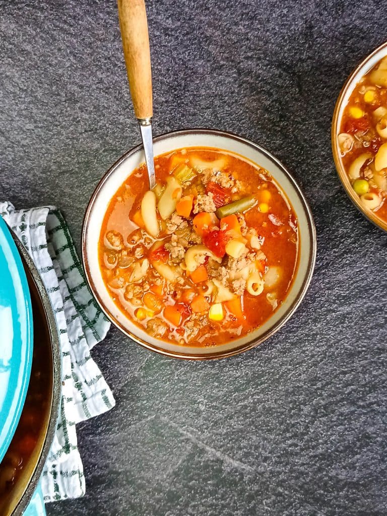 Two bowls and a blue pot of Busy Day Soup on a black background with a tablecloth. The hearty soup is made with elbow macaroni, ground beef, canned tomatoes, onions, celery, mixed vegetables, and beef stock. The hero bowl in the center has a spoon, showcasing the comforting, easy Busy Day Soup recipe.