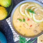 An overhead image of a bowl. In it there is a lemon and rice soup and some of the props include lemons and squeezed lemon, herbs and a pot of the soup beside the bowl and the bowl is on top of a green and white table cloth. The image has a black background.