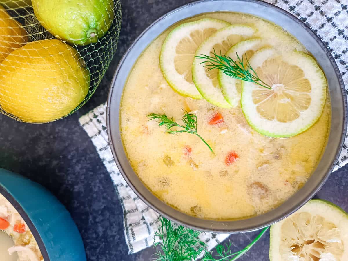 An overhead image of a bowl. In it there is a lemon and rice soup and some of the props include lemons and squeezed lemon, herbs and a pot of the soup beside the bowl and the bowl is on top of a green and white table cloth. The image has a black background.