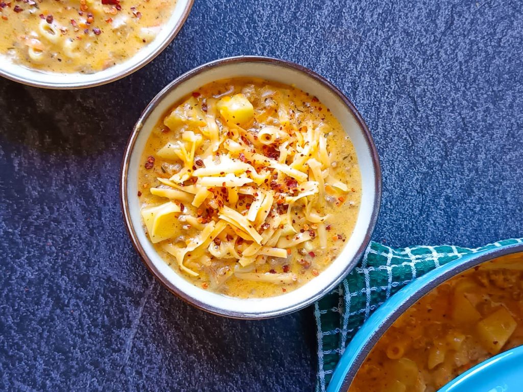 Two bowls of cheeseburger macaroni soup, the other one is sprinkled with chili flakes, the other one with grated cheese with a blue pot beside the two bowls of the soup. The pot is on top of a green table cloth and this picture has a black marble background.