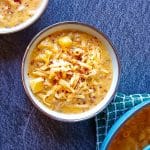 Two bowls of cheeseburger macaroni soup, the other one is sprinkled with chili flakes, the other one with grated cheese with a blue pot beside the two bowls of the soup. The pot is on top of a green table cloth and this picture has a black marble background.