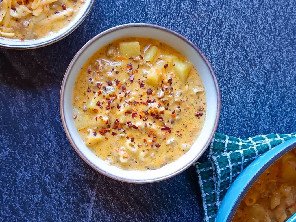 Two bowls of cheeseburger macaroni soup, the other one is sprinkled with chili flakes, the other one with grated cheese with a blue pot beside the two bowls of the soup. The pot is on top of a green table cloth and this picture has a black marble background.