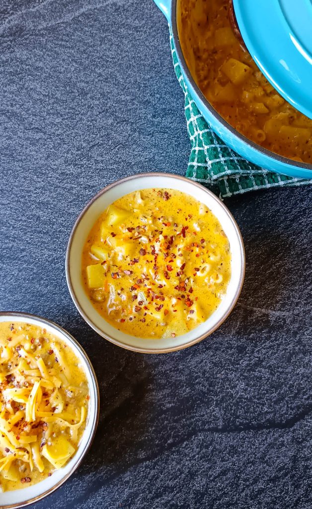 Two bowls of cheeseburger macaroni soup, the other one is sprinkled with chili flakes, the other one with grated cheese with a blue pot beside the two bowls of the soup. The pot is on top of a green table cloth and this picture has a black marble background.
