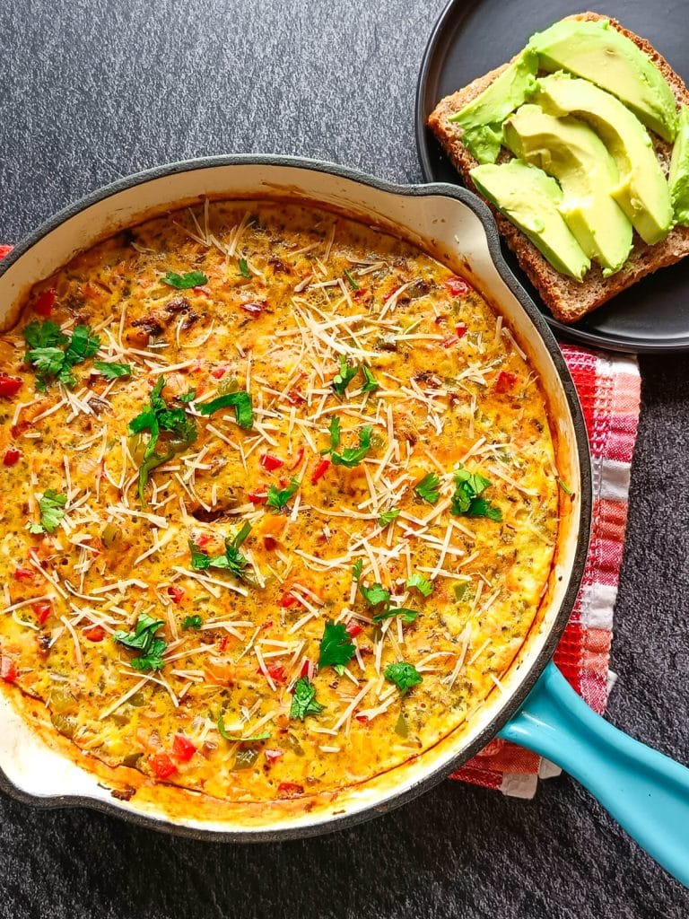 An overhead image of a blue cast iron pan with egg stew in it and beside it there is a black plate with brown bread topped with avocados. All this is on a black background and the pan is on top of an orange and red tablecloth.