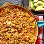 An overhead image of a blue cast iron pan with egg stew in it and beside it there is a black plate with brown bread topped with avocados. All this is on a black background and the pan is on top of an orange and red tablecloth.