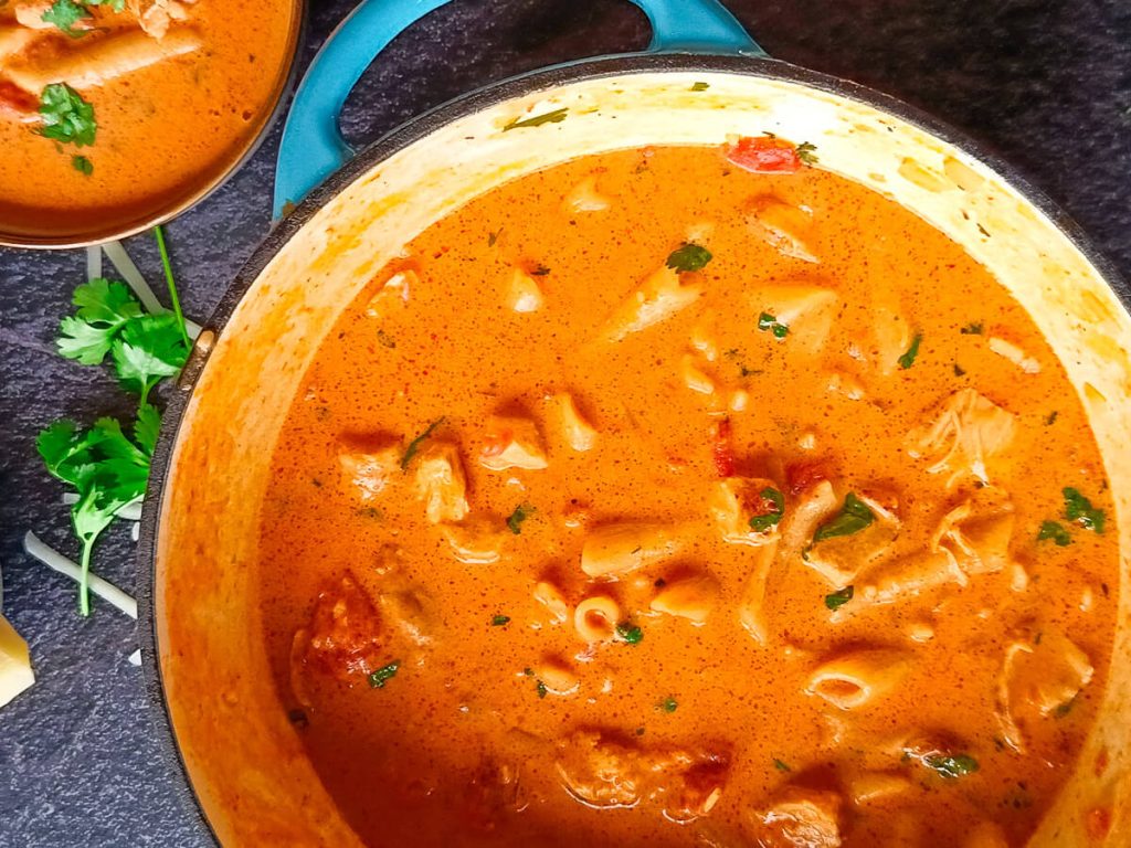 An image of a chicken parmesan soup in a cast-iron pot with a serving bowl next to it and other leftover ingredients, all on a black background.