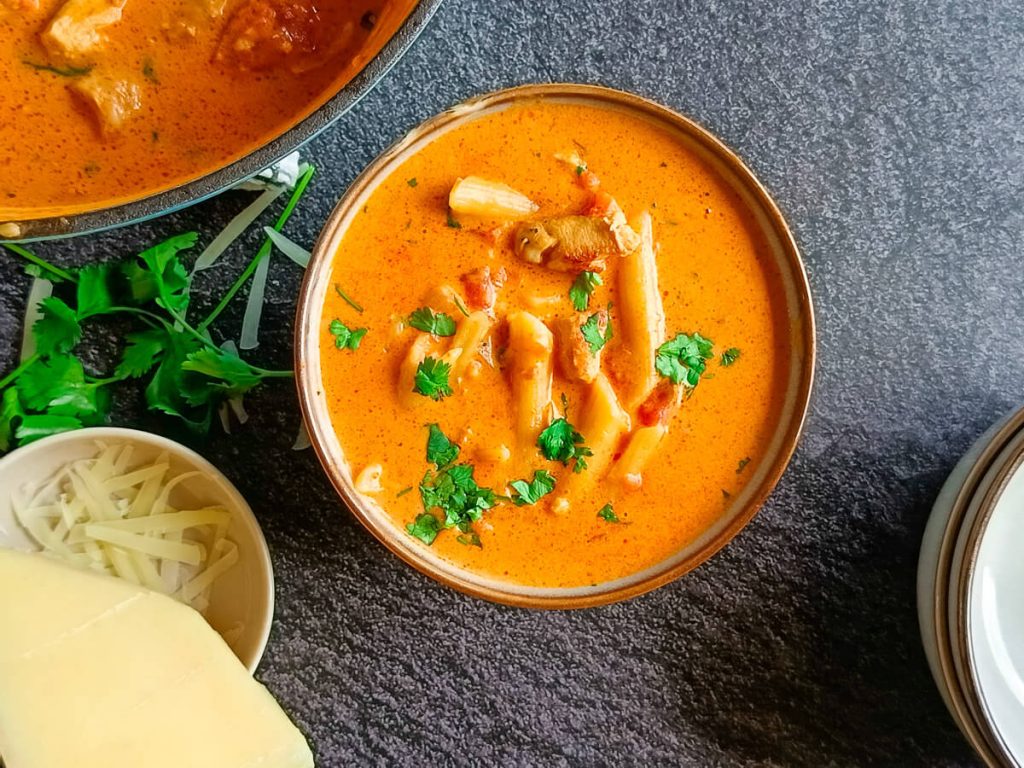 An overhead shot of a chicken parm soup in a bowl garnished with fresh coriander. There is a pot of the soup next to the bowl with more serving bowls and leftover ingredients all on a black background