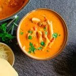 An overhead shot of a chicken parm soup in a bowl garnished with fresh coriander. There is a pot of the soup next to the bowl with more serving bowls and leftover ingredients all on a black background