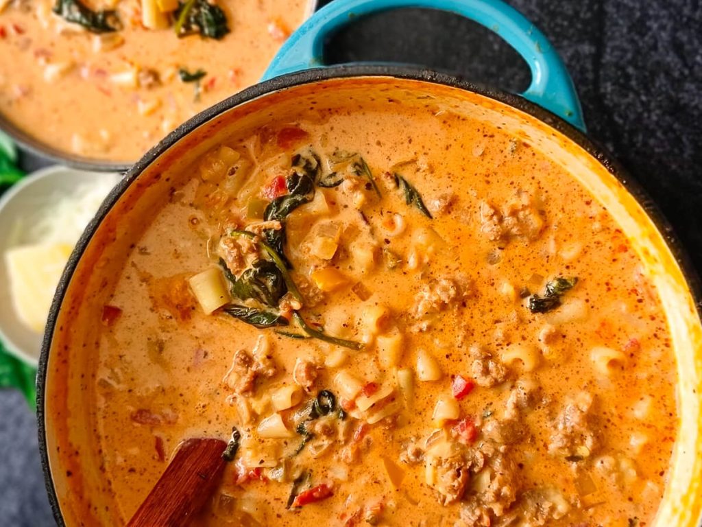 An image of parmesan Italian Sausage soup in a blue cast-iron pot with a wooden serving spoon inside it on a black background.