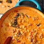 An image of parmesan Italian Sausage soup in a blue cast-iron pot with a wooden serving spoon inside it on a black background.