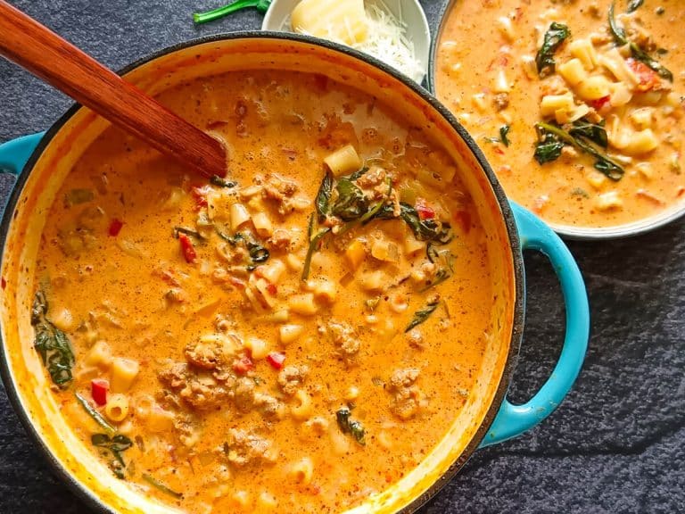 An image of Parmesan Italian Sausage soup in a blue cast-iron pot with a wooden serving spoon inside it and a bowl of soup beside it with other leftover ingredients on a black background.