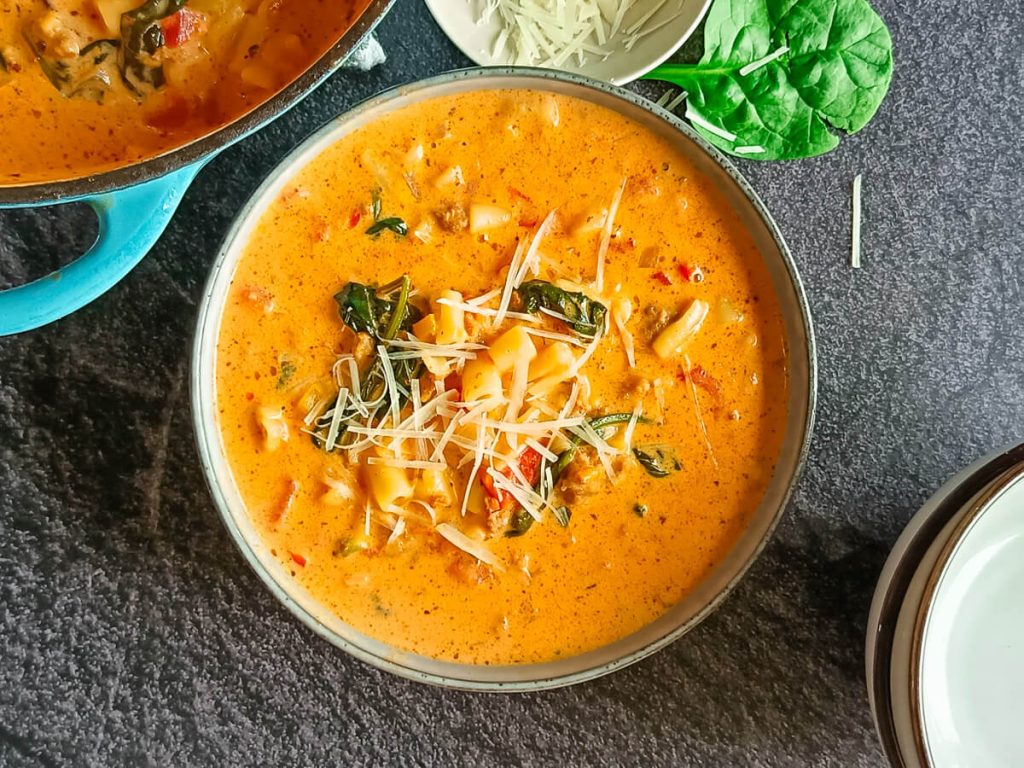 An image of parmesan Italian Sausage soup in a bowl with a blue cast-iron pot beside it on a black background.