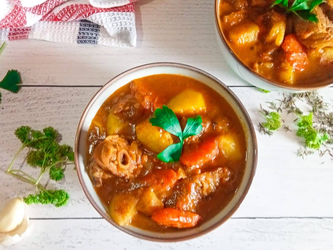 an overhead image of two bowls with turkey neck bone soup in them with fresh herbs scattered around the bowls and a table cloth in the corner.